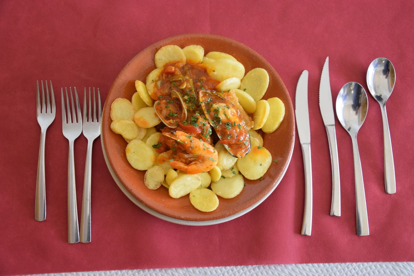 Bacalhau à marinheiro servido à mesa, com talheres sobre a toalha grená