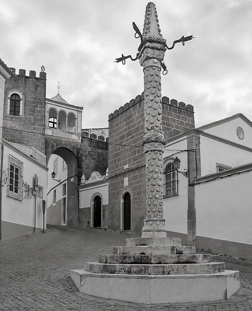 O pelourinho de Elvas no Largo de Santa Clara, com o arco e as torres da antiga cerca por trás