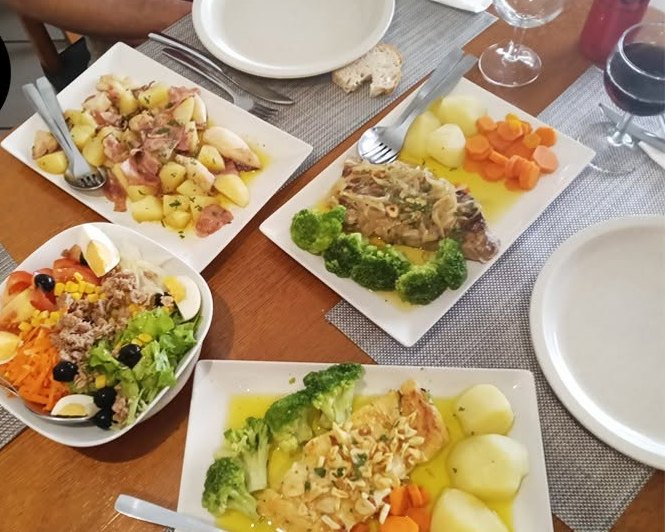 Uma mesa servida, vista de cima: saladas, bacalhau com batata e legumes, e pratos de carne