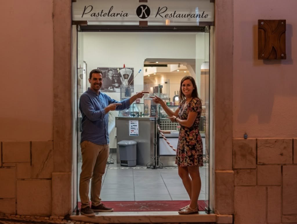 À porta do Kudissama, à noite, sob o letreiro Pastelaria Restaurante, com a sala iluminada por trás